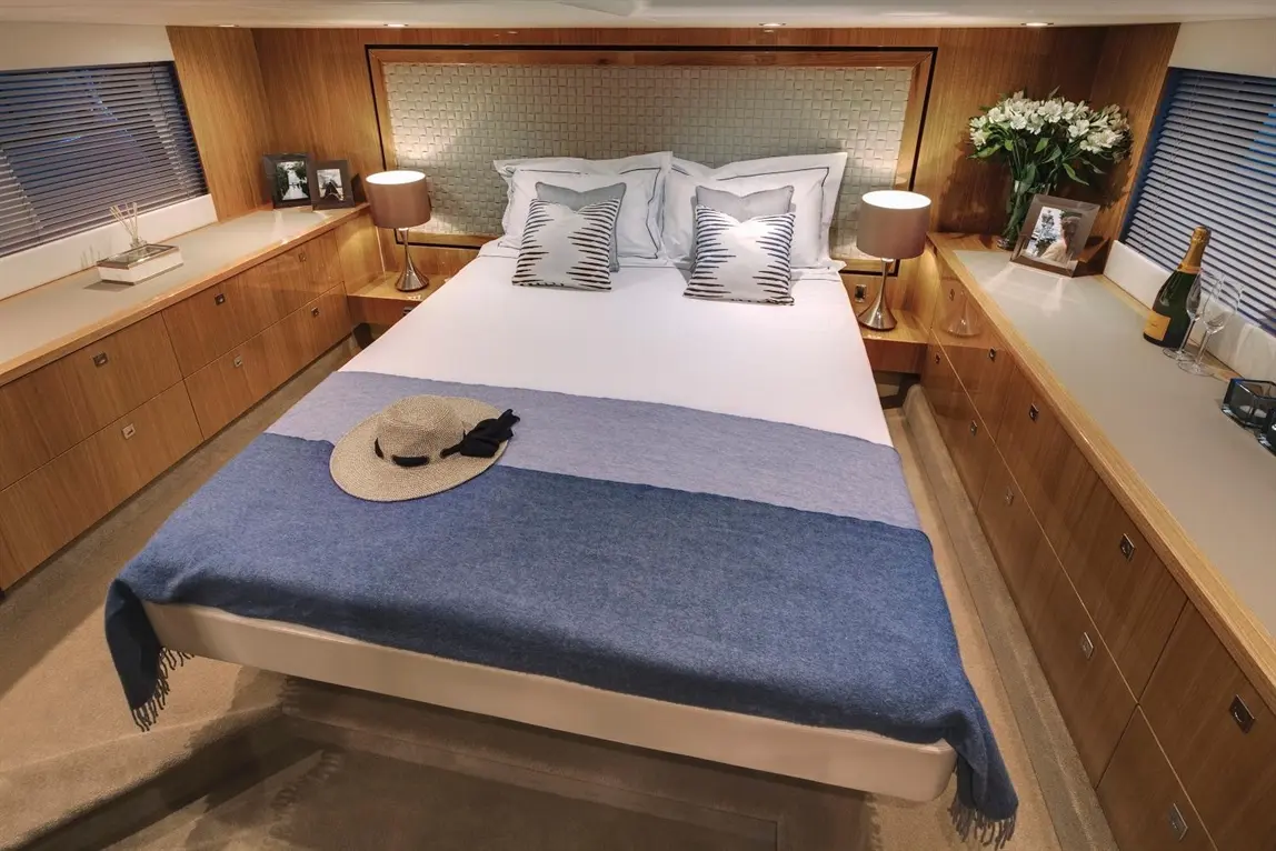 A stylish yacht bedroom featuring a double bed with blue throws, pillows, and a straw hat, alongside elegant furnishings.